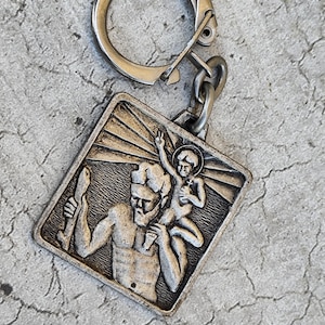 Op de afbeelding: Een zilverkleurige metalen sleutelhanger met een vierkante hanger met een reliëf van een bebaarde figuur die een klein kind op de schouders draagt. De figuur houdt een staf vast en het kind heeft een aureool. Het metaal heeft een antieke afwerking.
