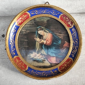 May include: Round, vintage religious artwork in a gold frame with blue and red accents. The image depicts a woman in a blue and pink robe, kneeling over a baby. The frame has a decorative design.