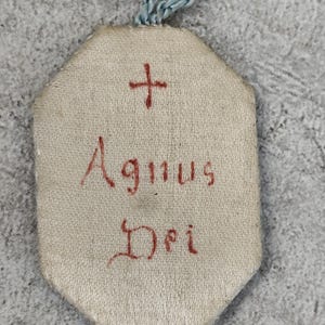 May include: An octagonal beige fabric pendant featuring a small blue cord loop at the top. The front surface displays a red embroidered cross above the red stitched text "Agnus Dei" centered on the fabric, with visible thread texture.