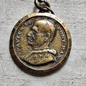 Pode incluir: Uma medalha de bronze com um perfil do Papa Paulo VI na frente. A medalha tem a inscrição "PAUS VI PONTIF MAX" ao redor da borda.