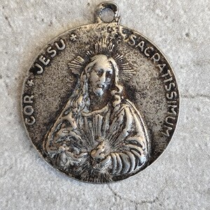 Peut inclure: Un pendentif religieux circulaire argenté avec l'inscription "COR JESU SACRATISSIMUM". Le pendentif présente un relief détaillé d'une figure avec un halo et une boucle pour suspendre.
