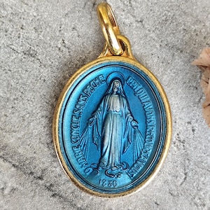 May include: An oval-shaped pendant featuring a gold-toned metal frame and a vibrant blue center. The center depicts a figure with outstretched arms, surrounded by the text O MARIE CONCUE SANS PECHE PRIEZ POUR NOUS QUI AVONS RECOURS A VOUS and 1830.
