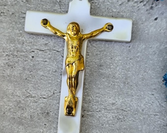 May include: A white crucifix with a gold-colored Jesus figure. The cross has a pearlescent finish, and the figure is detailed with outstretched arms. The crucifix is designed with a small hole at the top for hanging.