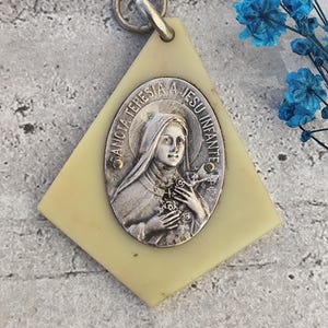 Op de afbeelding: Een witte plastic hanger met een zilveren ovaal medaillon met een afbeelding van Heilige Theresia van Lisieux die een boeket rozen vasthoudt. Het medaillon is gegraveerd met de tekst "Santa Teresa a Jesu Infante".