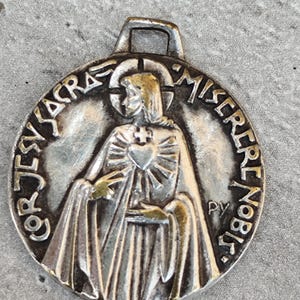 May include: A circular silver-toned metal pendant featuring a relief of a figure with a halo and a heart on the chest. The text COR JESU SACRA and MISERERE NOBIS is embossed around the edge, with the letters PY visible on the right side.