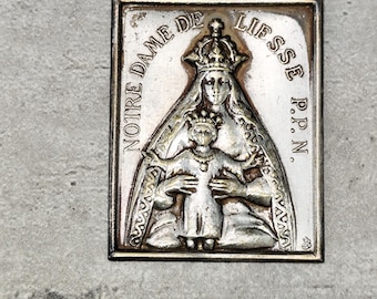 May include: A rectangular silver-colored religious pendant featuring a detailed relief of a crowned woman holding a child. The text "NOTRE DAME DE LIESSE P.P.N." is inscribed around the image. The pendant has a dark border and is set against a gray background.