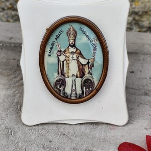 May include: A white, scalloped-edged, decorative plaque with an oval, framed image of a religious figure. The image features a man in ornate robes, holding a staff and a cross. Text on the frame reads "S. MAURO ABATE" and "VIAGRANDE (CT)".