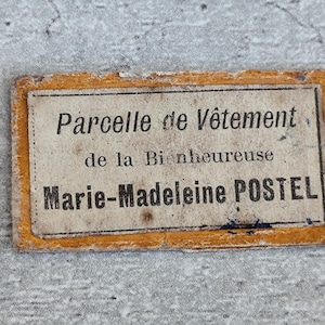 Peut inclure: Une étiquette rectangulaire en tissu avec un centre blanc cassé vieilli et une bordure effilochée jaune moutarde. Le texte indique : Parcelle de Vêtement de la Bienheureuse Marie-Madeleine POSTEL, imprimé en noir avec des signes d'usure.