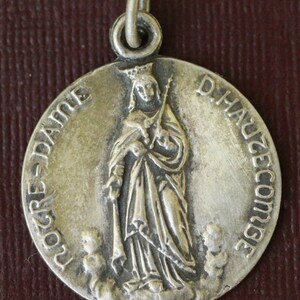 Könnte beinhalten: Silberner Anhänger mit einem religiösen Bild der Jungfrau Maria mit einer Krone und einem Stab. Die Inschrift lautet "Notre Dame D'Hautecourse".