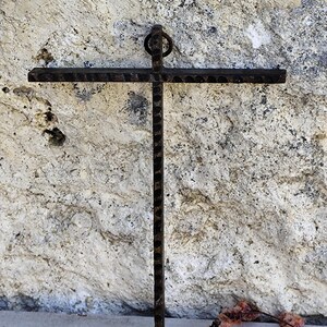 May include: A dark brown, wrought iron cross with a textured finish, set against a light-coloured stone wall. The cross has a ring at the top for hanging. The image includes dried flowers at the bottom, adding a natural element.