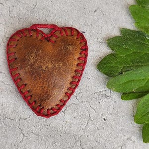 Può includere: Un ciondolo a forma di cuore in pelle fatto a mano con un bordo rosso cucito. Il ciondolo è di colore marrone caldo con una superficie strutturata. Le cuciture rosse creano un contorno contrastante. In alto c'è un anello di filo rosso per appenderlo.