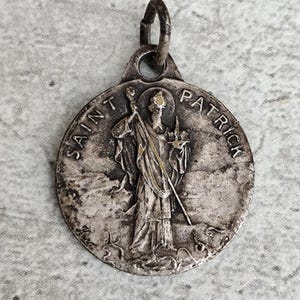 Può includere: Medaglia di San Patrizio in argento con una dettagliata raffigurazione di San Patrizio che tiene un bastone e una croce. Le parole "SAINT PATRICK" sono incise attorno al bordo del ciondolo circolare. La medaglia ha un anello per appenderla.