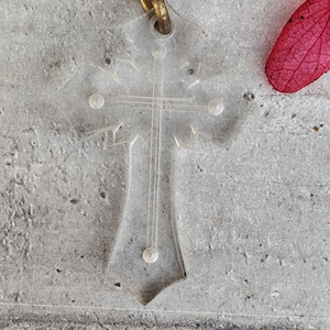 Puede incluir: Un colgante transparente en forma de cruz con una anilla dorada. El colgante presenta un diseño de cruz con cuatro pequeños adornos redondos. El fondo es una superficie gris texturizada, con un pétalo rojo en la esquina superior derecha.