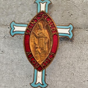 May include: A vintage religious cross-shaped pin with a central oval design. The oval features an illustration of a religious figure and the words "SAINT CLAUDE ALOURDES". The cross arms are light blue with white and red accents.