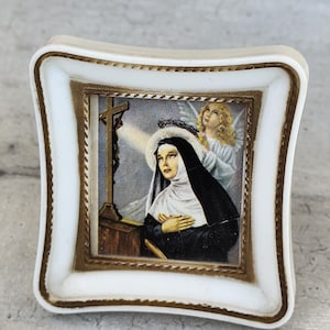 Può includere: Piccola immagine religiosa quadrata, bordata di bianco e oro. L'immagine raffigura una suora con un angelo sullo sfondo. La suora indossa un abito nero e un velo bianco. Una croce dorata è visibile sul lato sinistro.