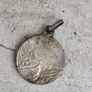 May include: A round, silver-toned pendant with a detailed relief design. The pendant features a depiction of a car and a landscape. The bail is attached at the top. The pendant has an aged, vintage appearance.