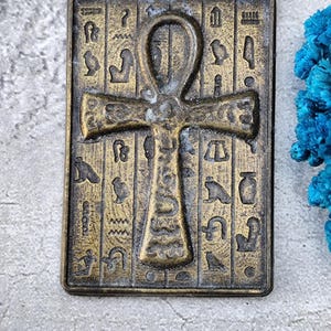 Può includere: Un ciondolo rettangolare color bronzo con un simbolo Ankh al centro, con incisioni geroglifiche. Il ciondolo ha un aspetto invecchiato, che suggerisce un'estetica antica o vintage. Lo sfondo è una superficie grigia strutturata.
