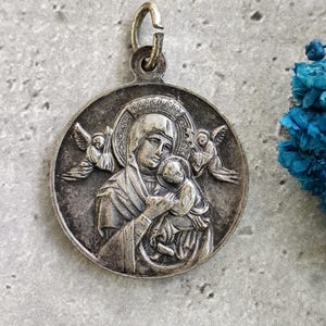 Pode incluir: Pingente prateado com uma representação da Virgem Maria segurando o menino Jesus. O pingente é circular e tem um laço na parte superior para pendurar.