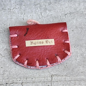 Può includere: Piccola custodia in pelle rossa con cuciture rosa sui bordi. La custodia ha un piccolo anello in alto e un'etichetta rettangolare con la scritta "Agnus Dei". Su una superficie grigia.