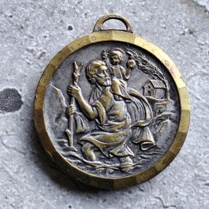 Pode incluir: Pingente circular em bronze antigo com um relevo detalhado de um santo carregando uma criança, com um bastão. O pingente tem uma forma circular com uma argola na parte superior para pendurar. A palavra "ITALY" é visível.
