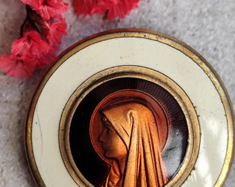 Könnte beinhalten: Ein runder religiöser Anhänger mit goldfarbenem Rand und weißem Emaille-Rand. In der Mitte befindet sich ein kupferfarbenes Profil einer Frau mit Heiligenschein. Rote Blumen sind im Hintergrund.