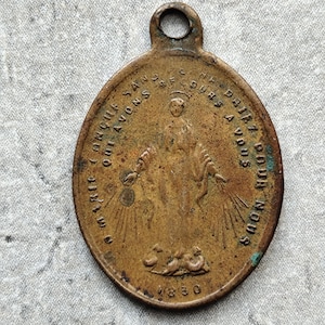 Peut inclure: Pendentif ovale en bronze ancien avec imagerie religieuse. Le pendentif présente une image en relief d'une figure aux bras tendus, entourée de texte. L'année "1830" est inscrite en bas. Il possède une boucle en haut pour l'accrocher.