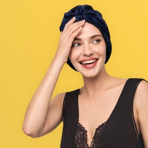 May include: A woman with a bright smile is wearing a navy blue satin hair wrap. She is wearing a black sleeveless top with a lace trim.