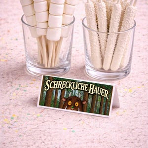 May include: Two clear glasses filled with treats. One holds marshmallow skewers, the other white chocolate-covered pretzel sticks. A small card with the text "SCHRECKLICHE HAUER" and a cartoon forest scene is in front.