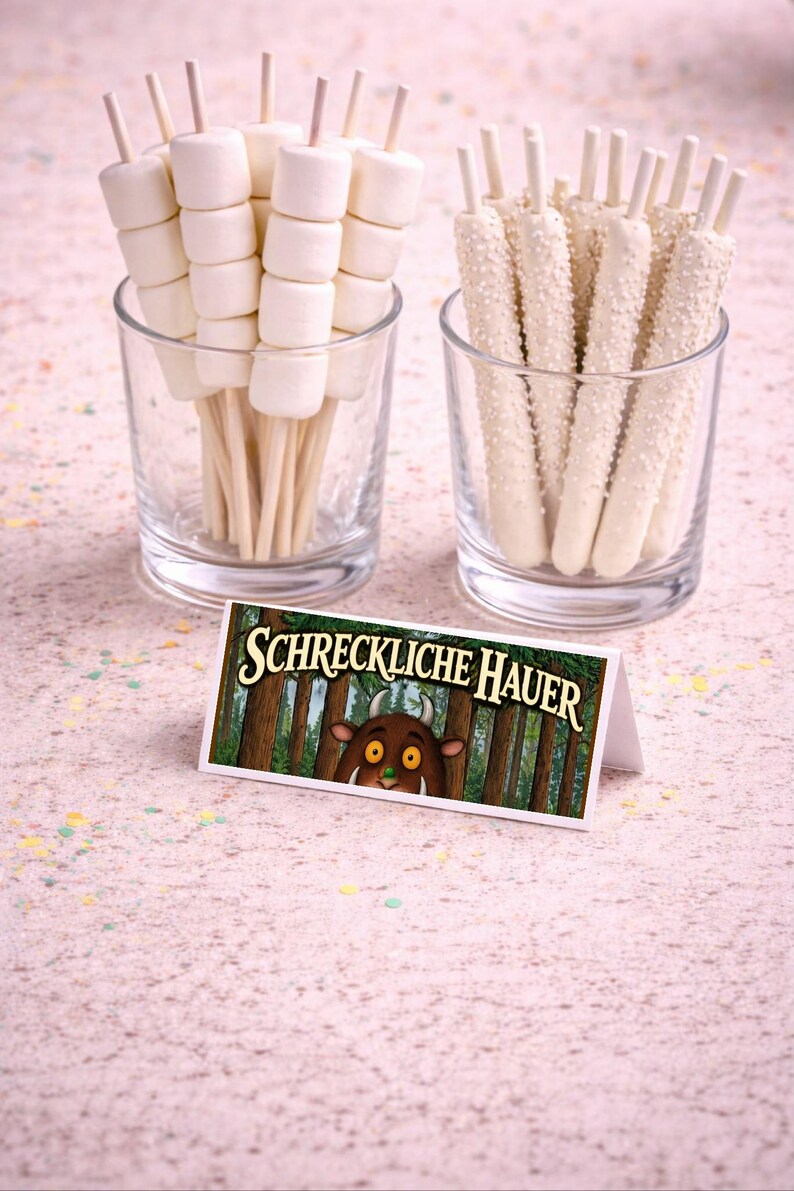 May include: Two clear glasses filled with treats. One holds marshmallow skewers, the other white chocolate-covered pretzel sticks. A small card with the text "SCHRECKLICHE HAUER" and a cartoon forest scene is in front.