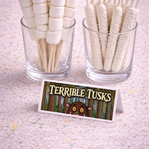 May include: Two clear glasses with treats. One glass holds marshmallow skewers, the other holds white chocolate-covered pretzel sticks. A small sign reads "TERRIBLE TUSKS" with a cartoon illustration.