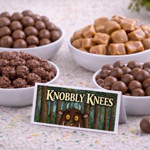 May include: Four white bowls filled with different candies: chocolate malt balls, caramel cubes, and chocolate-covered clusters. A sign reads "Knobbly Knees" with a forest illustration and a cartoon character.