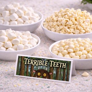 May include: Four white bowls filled with different snacks, including marshmallows, popcorn, and round, white candies. A sign in front reads "TERRIBLE TEETH" with a cartoon monster illustration, suggesting a themed food display.