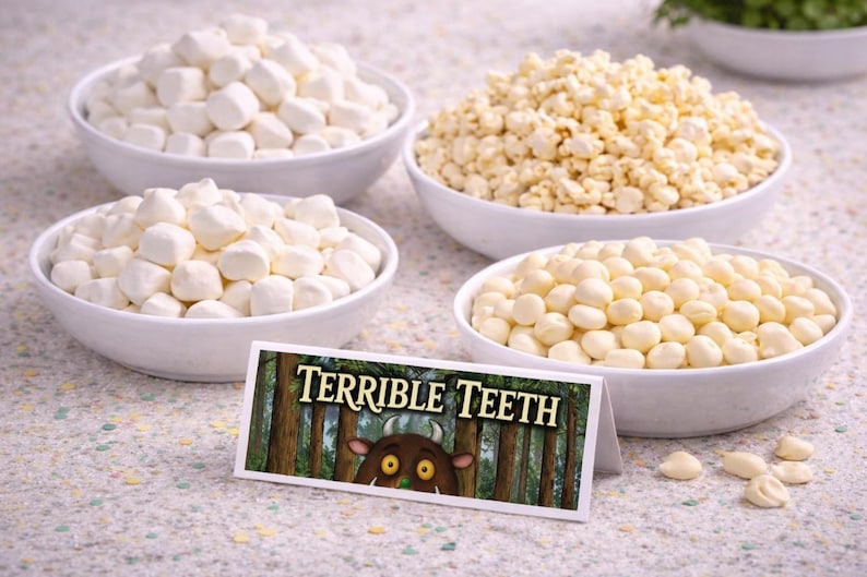 May include: Four white bowls filled with different snacks, including marshmallows, popcorn, and round, white candies. A sign in front reads "TERRIBLE TEETH" with a cartoon monster illustration, suggesting a themed food display.