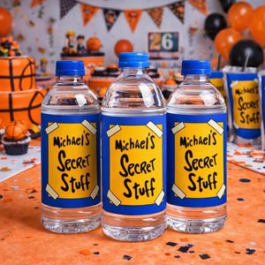 May include: Three clear water bottles with blue caps and labels that read "Michael's Secret Stuff" in black lettering on a yellow background. The bottles are on an orange surface, with party decorations in the background.