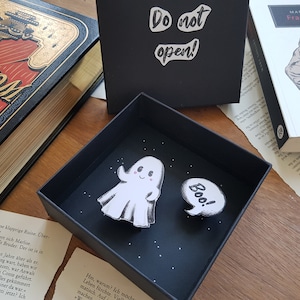 Könnte beinhalten: Eine schwarze Box mit einem weißen Geist und einer Sprechblase, die "Boo!" sagt. Die Box ist geschlossen und hat die Aufschrift "Nicht öffnen!" auf der Oberseite.