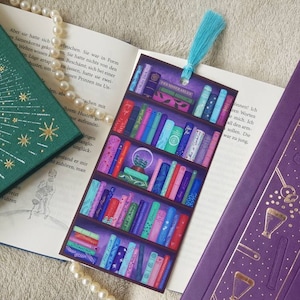 Könnte beinhalten: Nahaufnahme eines Buches mit einem Lesezeichen, das ein Bücherregal mit bunten Büchern zeigt. Das Lesezeichen ist aus Stoff und hat einen Quasten. Das Buch ist lila mit goldenen Akzenten und der Text "TRIS MINERARIES" ist auf dem Lesezeichen sichtbar.