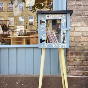May include: A blue wooden Little Free Library with a white floral design and a glass door filled with books. The library is on a wooden stand with four legs.