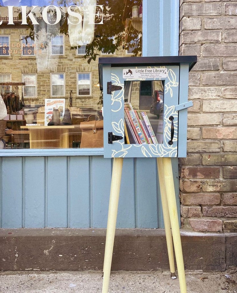 May include: A blue wooden Little Free Library with a white floral design and a glass door filled with books. The library is on a wooden stand with four legs.