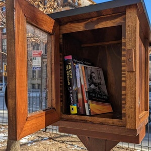 May include: A wooden book exchange box with a glass door open, revealing a shelf with several books. The books include "The City We Became" by N.K. Jemisin and "The Fire Next Time" by James Baldwin.