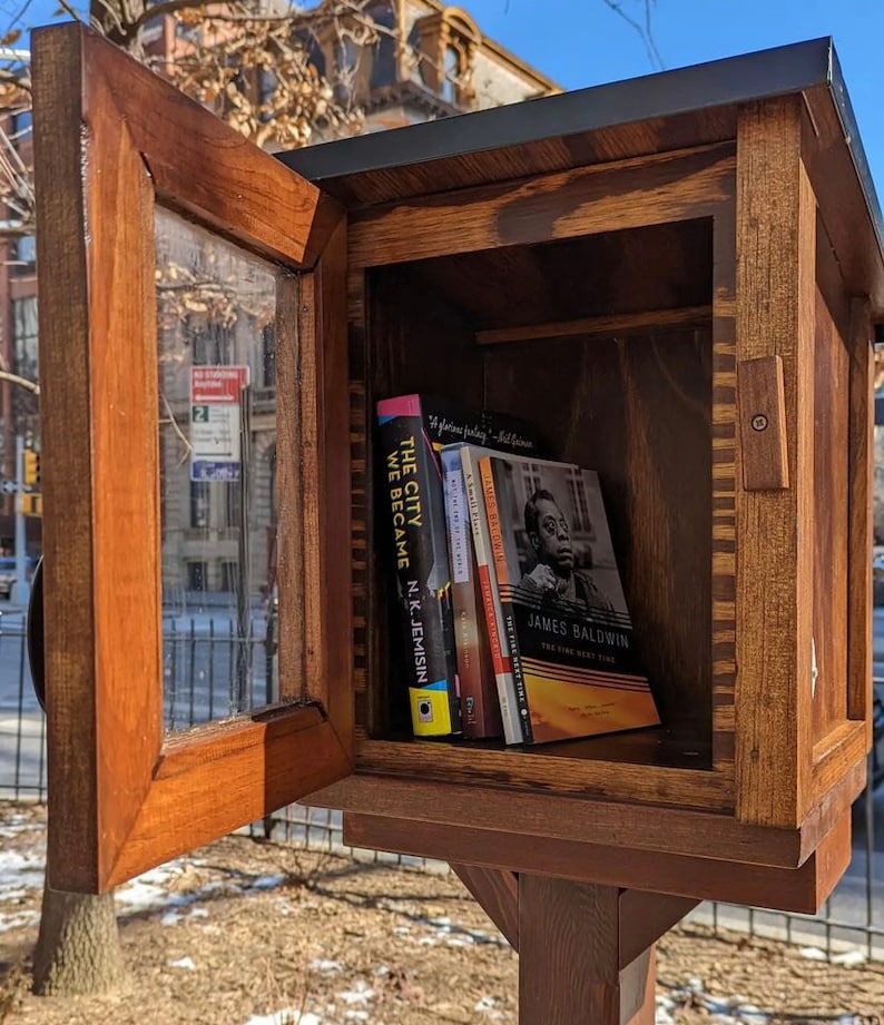 May include: A wooden book exchange box with a glass door open, revealing a shelf with several books. The books include "The City We Became" by N.K. Jemisin and "The Fire Next Time" by James Baldwin.