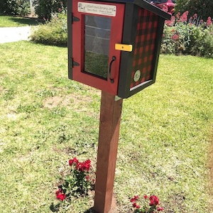 May include: A red and black checkered Little Free Library book exchange box with a glass door and a yellow latch. The box is mounted on a wooden post and surrounded by green grass and red flowers.