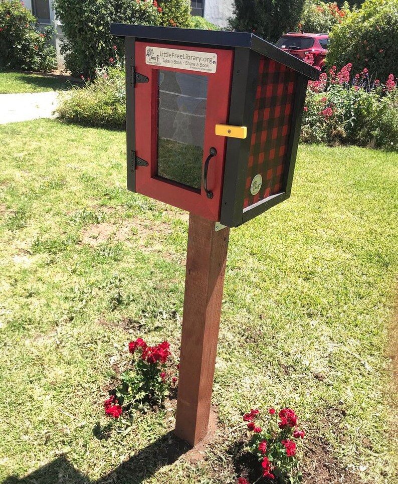May include: A red and black checkered Little Free Library book exchange box with a glass door and a yellow latch. The box is mounted on a wooden post and surrounded by green grass and red flowers.