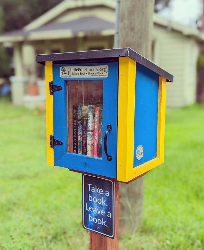 May include: A blue and yellow Little Free Library book exchange box with a sign that reads "Take a book. Leave a book." The box is filled with books and is located in a grassy area.