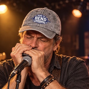 May include: A grey distressed baseball cap featuring the text BORN TO BEND in white and blue lettering. The cap has a curved brim and a structured crown, displaying a graphic of a harmonica and musical notes below the primary text.