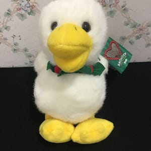 May include: A plush white duck with a yellow beak and feet, wearing a green and red bow tie. The duck has black eyes and a tag that reads "Snowden 1998". The stuffed animal is a cute and cuddly toy.