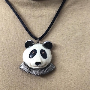 May include: A panda bear pendant necklace. The panda has a white face with black eyes and ears, and a silver ruffled collar. The pendant is suspended from a black cord.