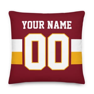 May include: A square throw pillow in deep maroon featuring the text YOUR NAME in white block letters above the large white numbers 00, which are outlined in yellow. A horizontal white and yellow stripe pattern runs across the centre of the pillow.