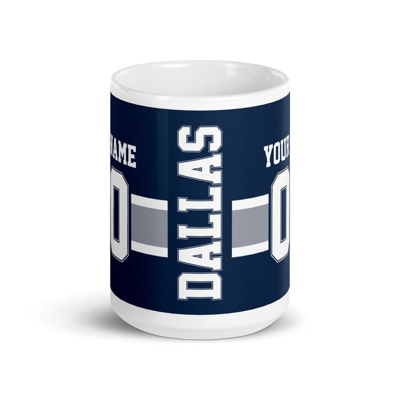 K&ouml;nnte beinhalten: Eine wei&szlig;e Keramiktasse mit einem blauen und grauen Design. Die Tasse zeigt das Wort "Dallas" in wei&szlig;er Schrift und eine wei&szlig;e Zahl "0" auf beiden Seiten. Der Text "Name" und "Your" ist ebenfalls auf der Tasse zu sehen.