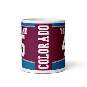 May include: White ceramic coffee mug with a burgundy, blue, and gray design. The mug features the word "COLORADO" in white lettering and a number "4" in gray lettering.