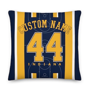 May include: A navy blue throw pillow with a yellow and white striped border. The center of the pillow features a basketball court design with the text "CUSTOM NAME" and the number "44" in yellow. The text "INDIANA" is also printed in yellow below the number.